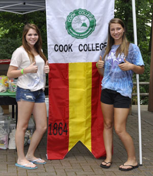 Two young alumni standing on either side of the Cook college gonfalon.